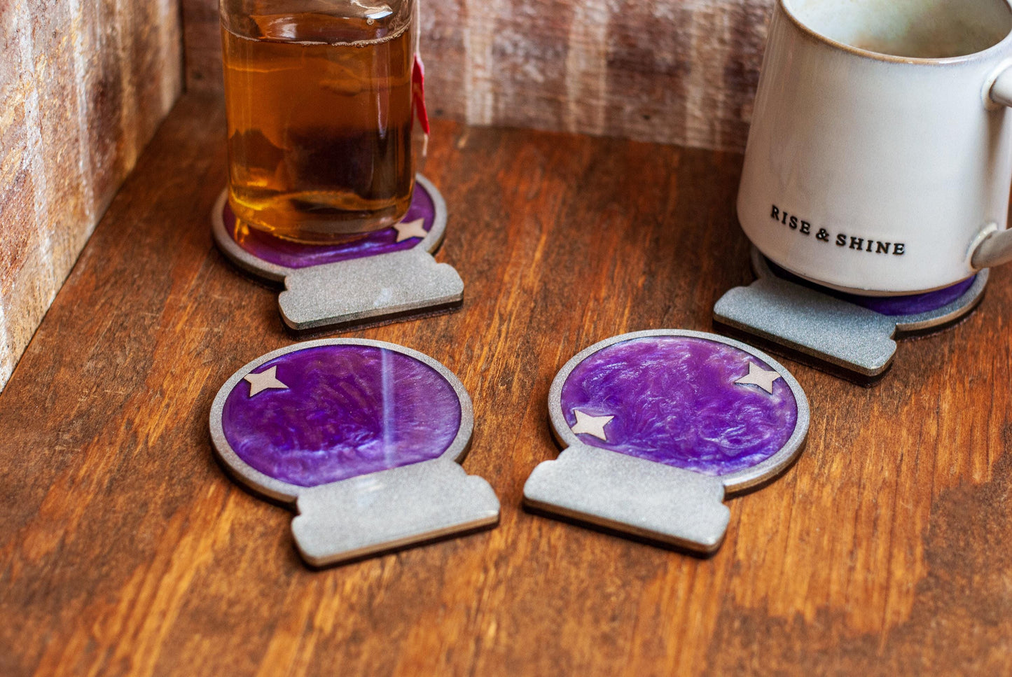 A set of four purple and silver mystical celestial-themed resin crystal ball coasters on a wooden table and cup of tea