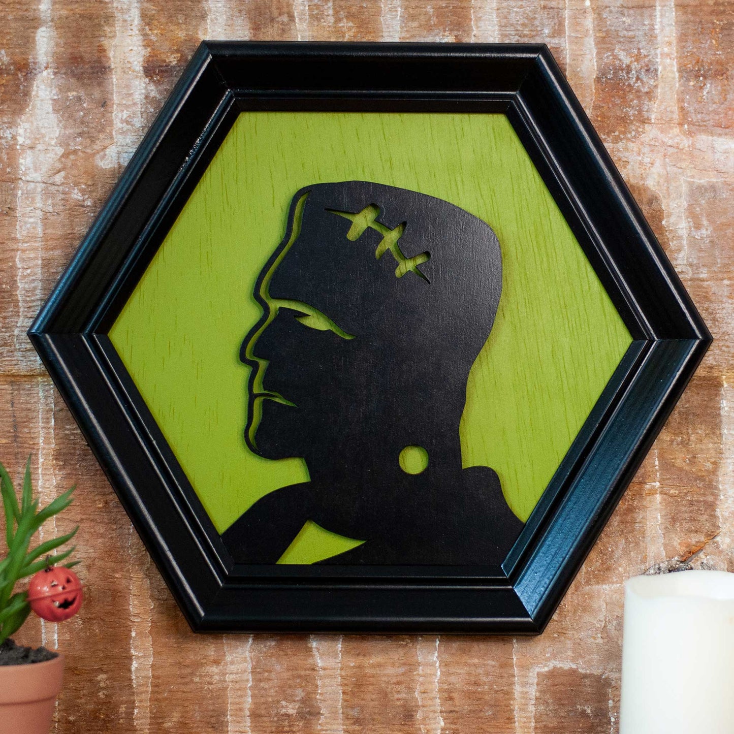Hexagonal frame with a black silhouette of a modern Frankenstein on a green background against a brick wall with plant and candles