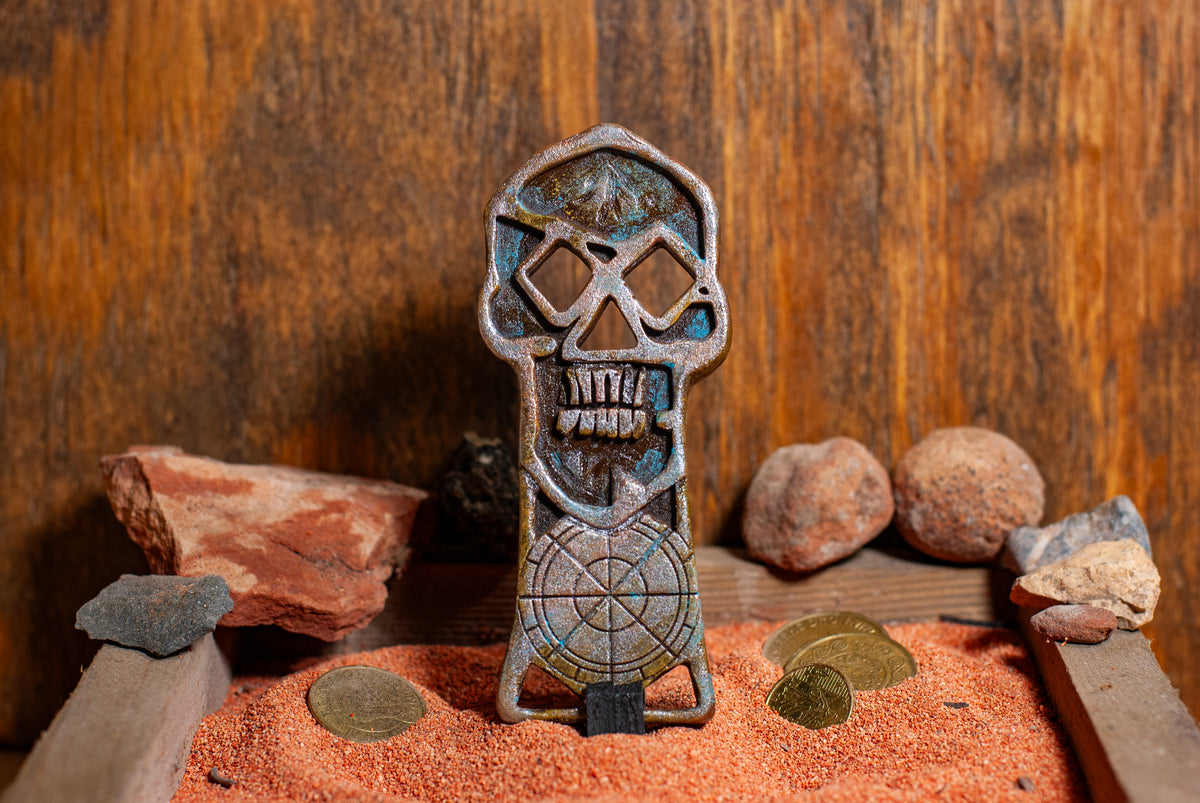 Front view of a copper bones skeleton key displayed in a wooden box of sand with gold coins in the background
