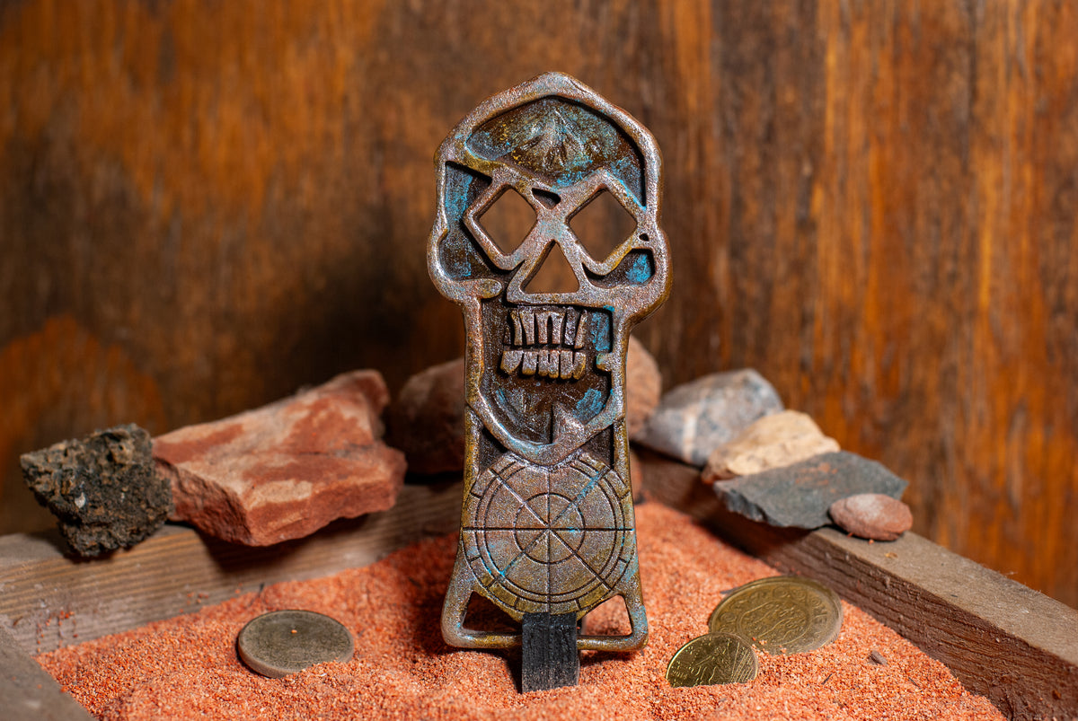 A copper bones skeleton key standing in a wooden box of sand with gold coins in the background