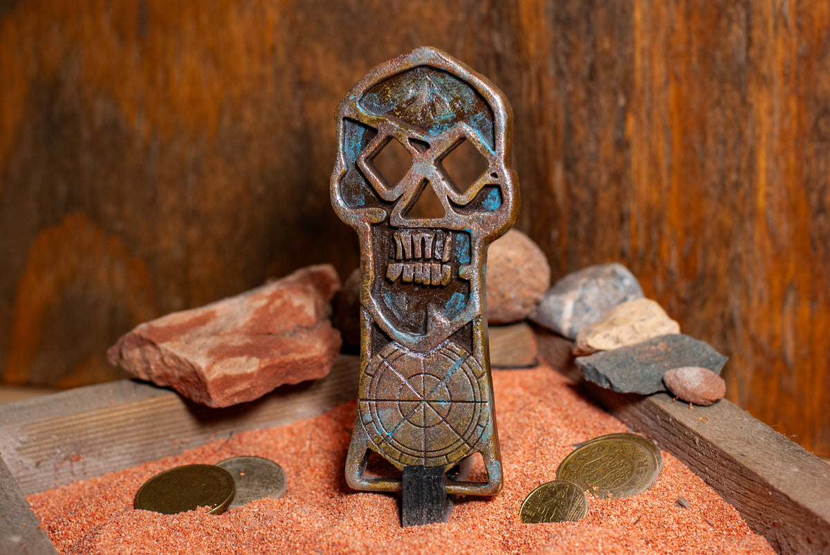 Side view of a copper bones skeleton key displayed in a wooden box of sand with gold coins in the background