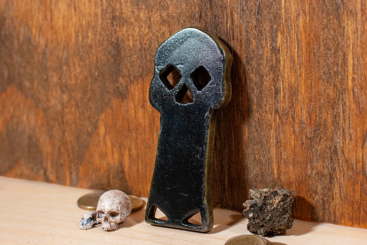 Back view of a copper bones skeleton key prop with skeleton bones and gold coins surrounding it and a wooden background