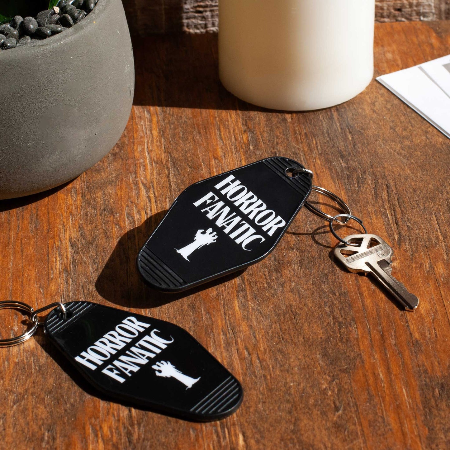 Two 'Horror Fanatic' keychains on a wooden surface with a plant and candle in the background.