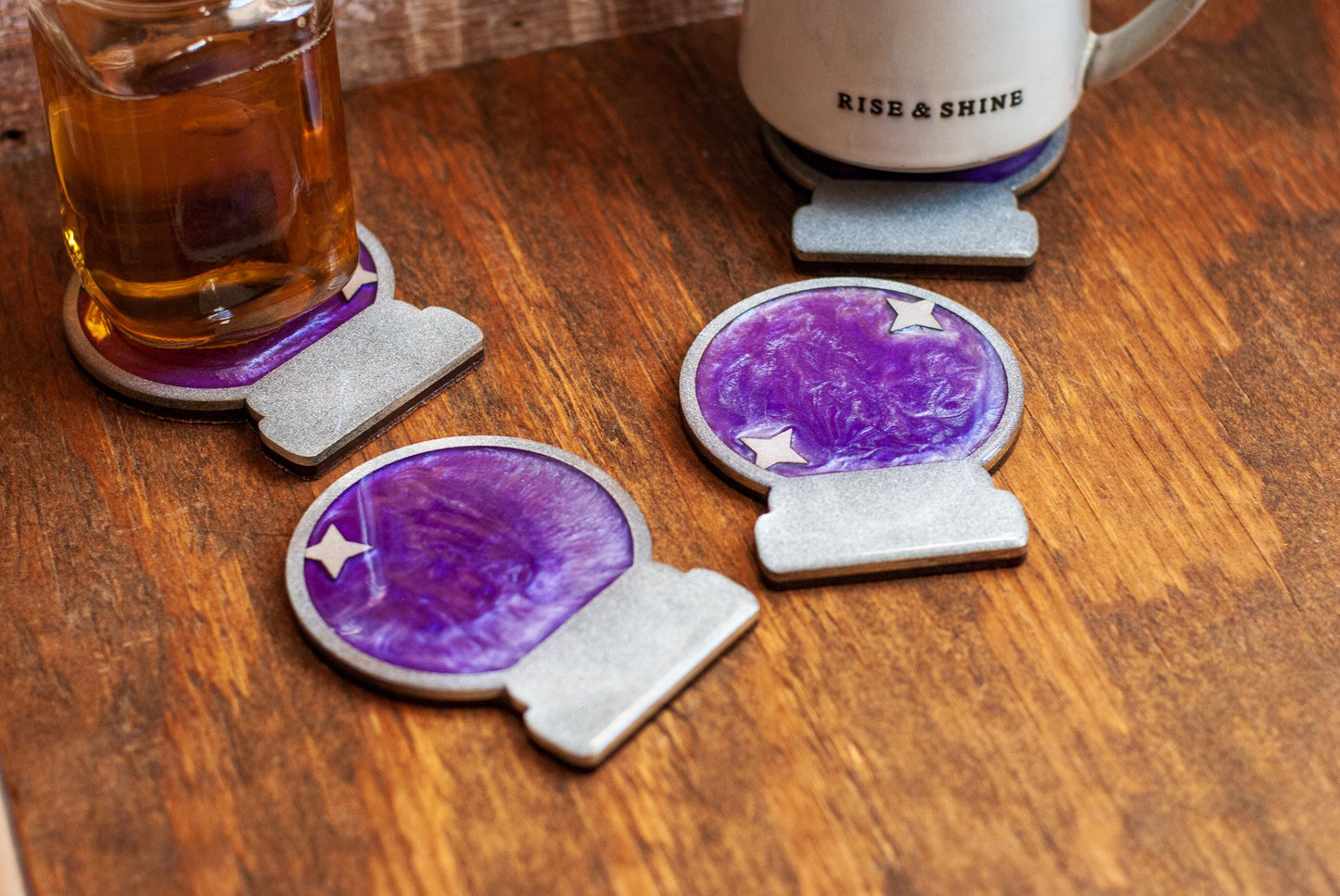 A set of two purple and silver mystical celestial-themed resin crystal ball coasters on a wooden table and cup of tea