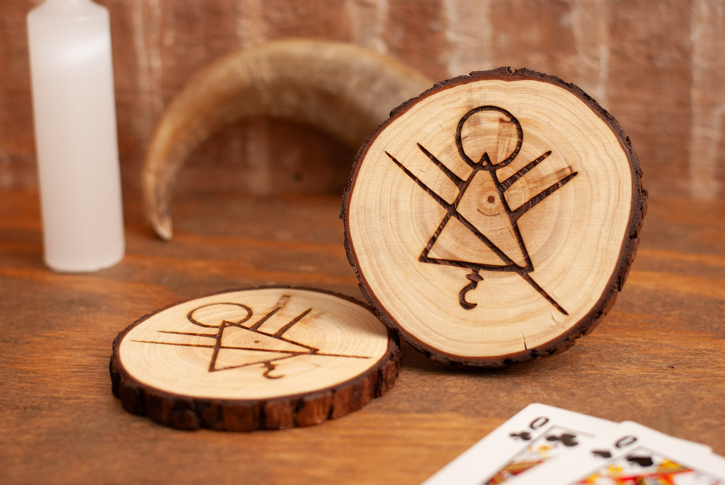 Wooden coasters with engraved rune symbols on a wooden surface with playing cards, candle, and horn in background.