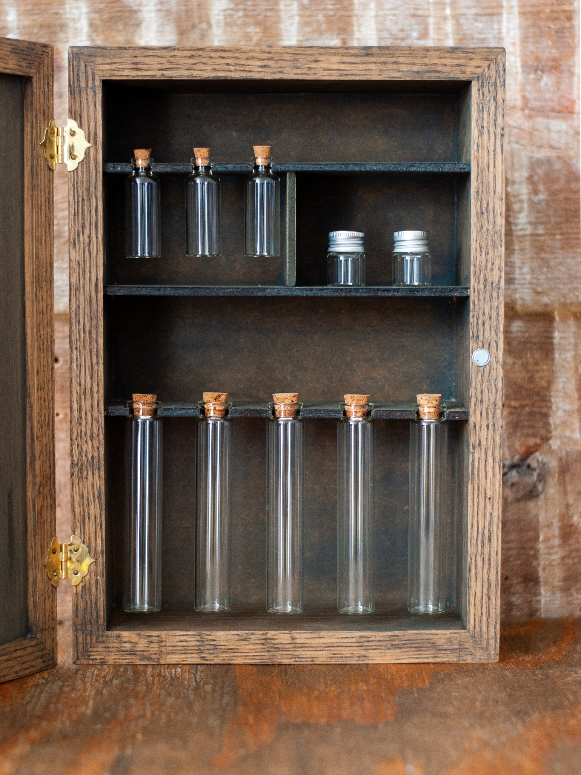 rustic wooden apothecary-style box with bronze and gold accents, glass vials and jars in various sizes with 'Apothecary Potions Elixirs' label on a wooden tabletop and brick background