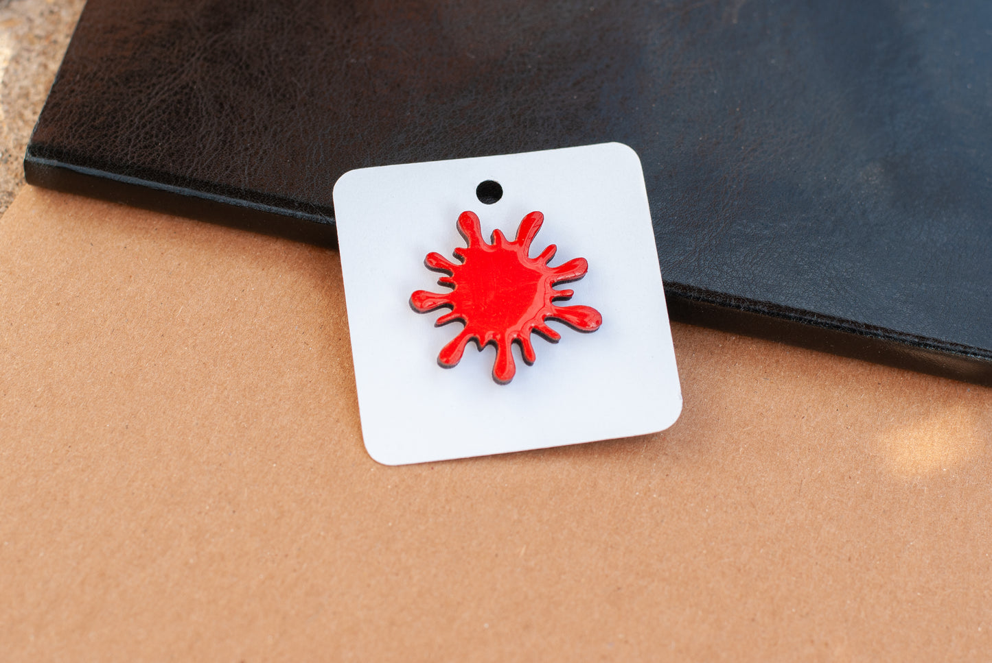 blood red splatter brooch pin on a white square card with a black border, placed on a brown surface.