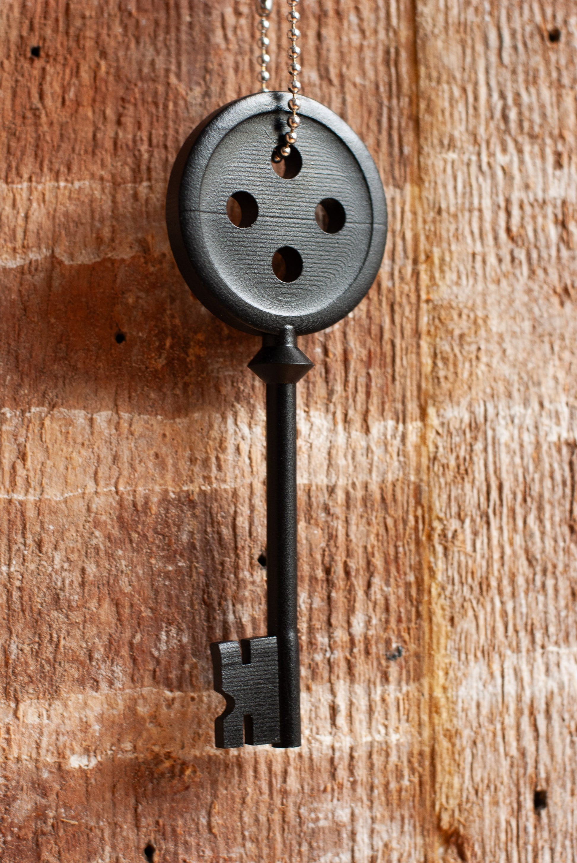Black Coraline inspired button key prop replica hanging from a silver ball chain on wooden table