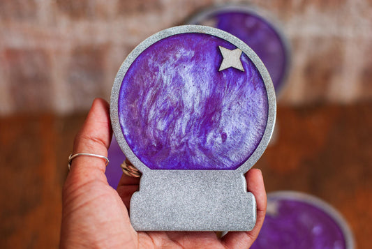 Hand holding a purple and silver mystical celestial-themed resin crystal ball coaster with a star on a wooden surface