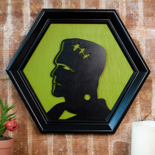 Hexagonal frame with a black silhouette of a modern Frankenstein on a green background against a brick wall with plant and candles