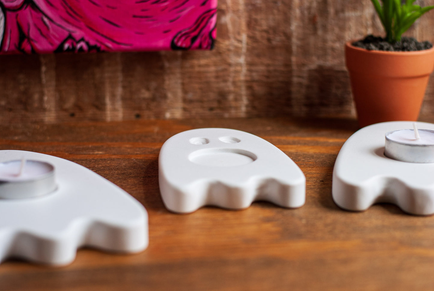 Three ghost-shaped candle holders on a wooden surface with a pink and black patterned object and a plant in the background.