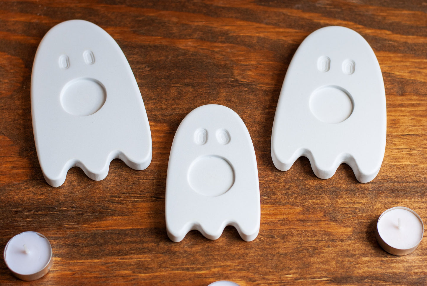 Three white ghost-shaped candle holders on a wooden surface with two small candles.