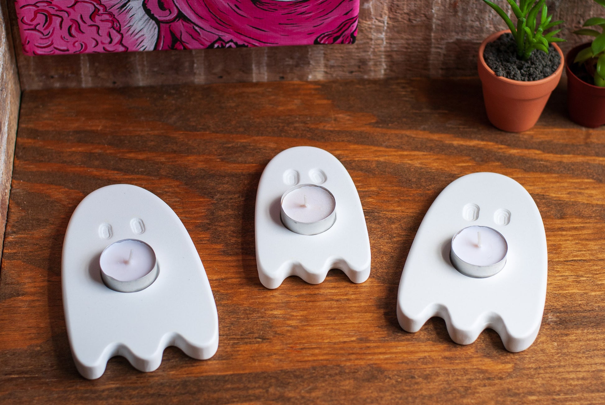 Three ghost-shaped candle holders on a wooden surface with plants in the background.
