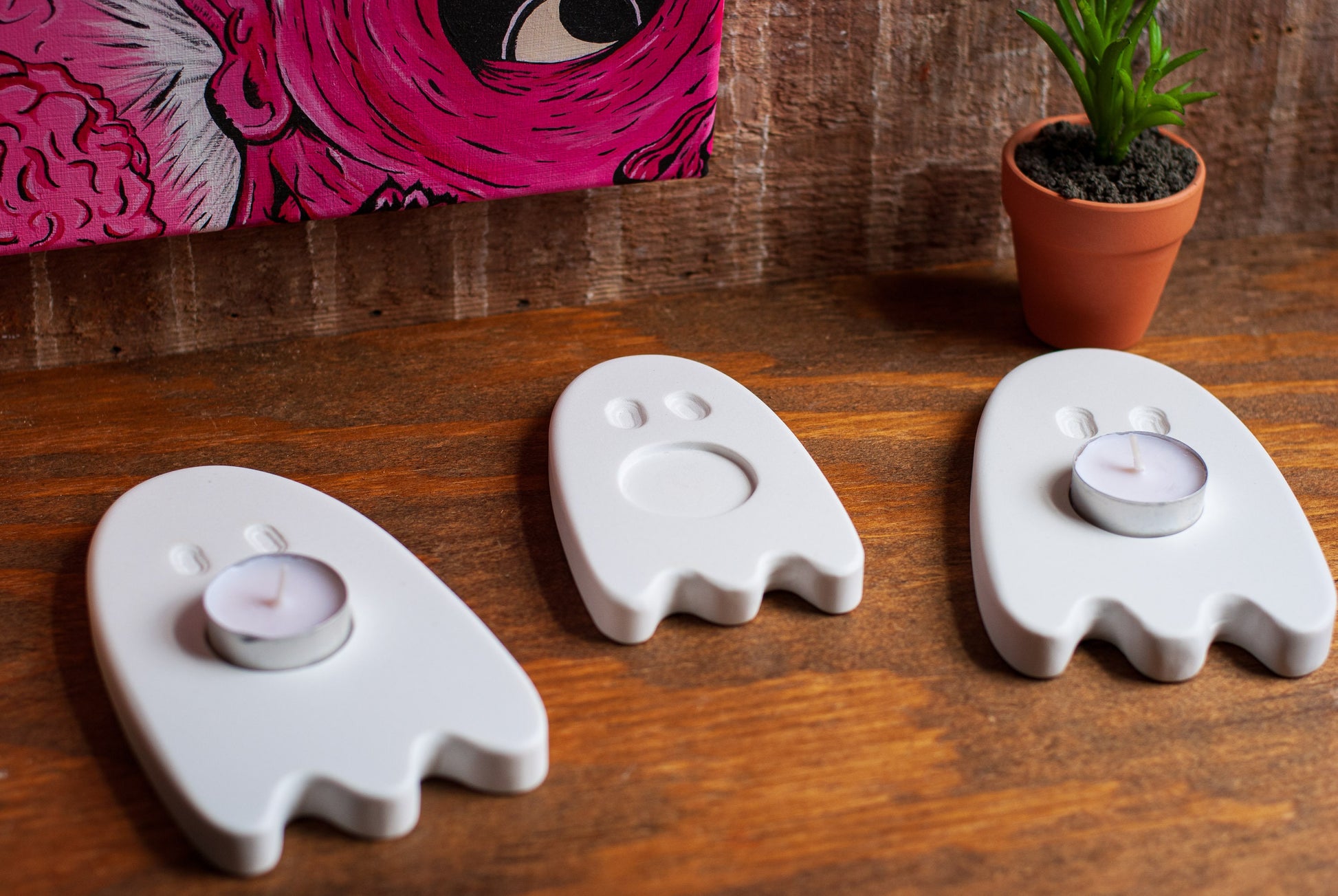 Three ghost-shaped candle holders on a wooden surface with a colorful painting and plant in the background.