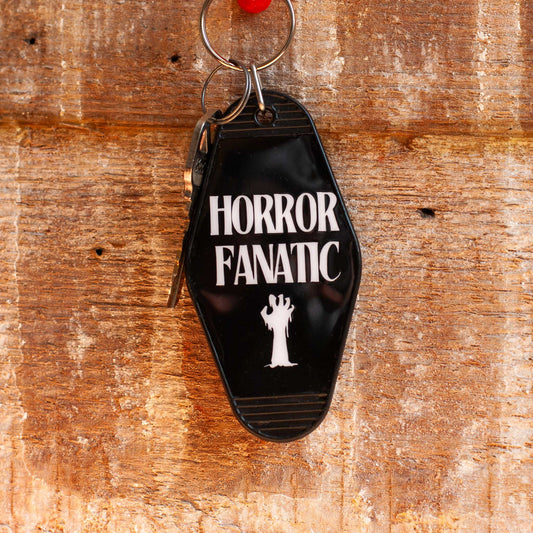 front view of black motel keychain with 'Horror Fanatic' text and logo hanging on hook on a wooden surface