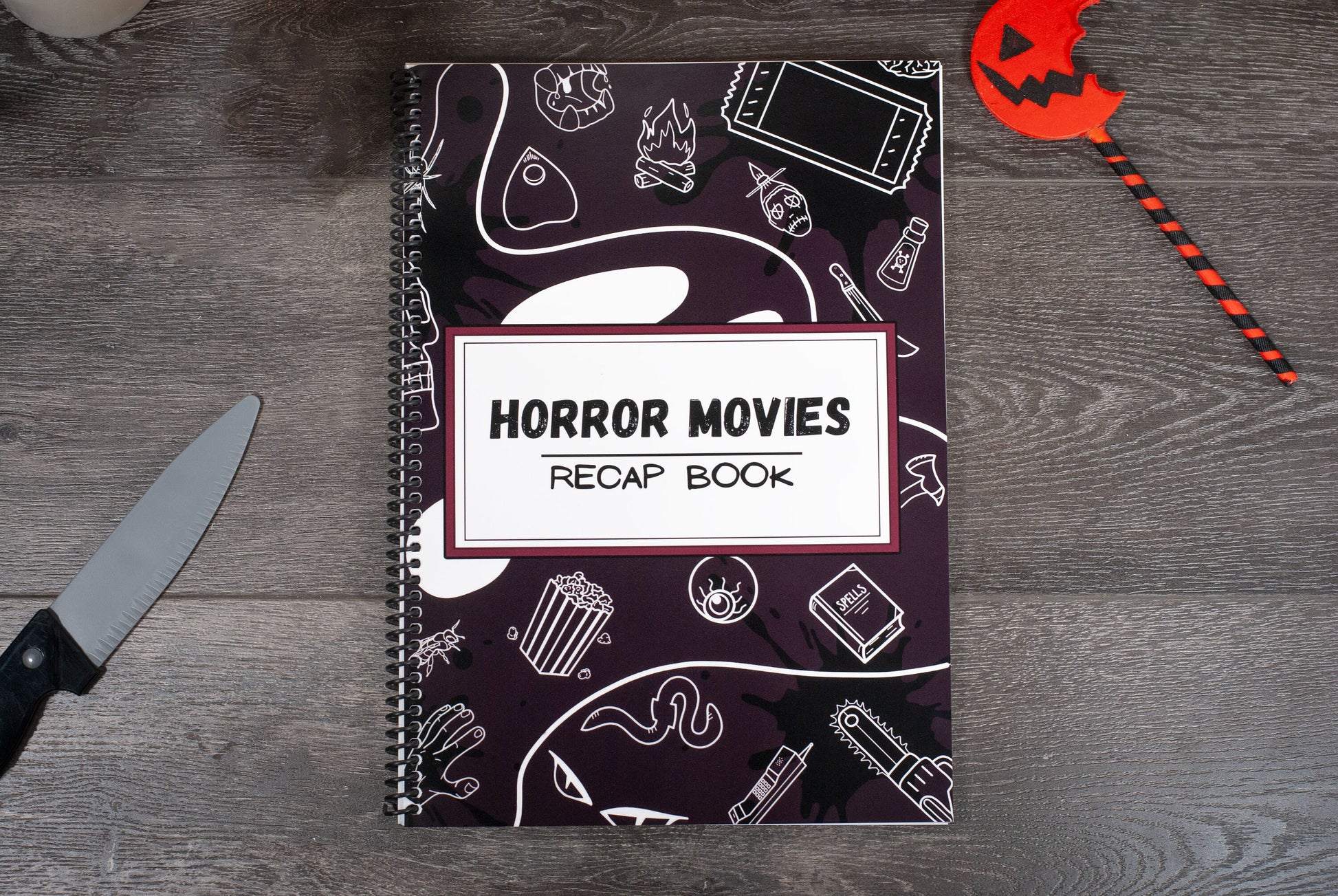 top down view of a spiral movie review journal titled Horror Movies Recap Book on wood table with pumpkin lollipop and Halloween decorations