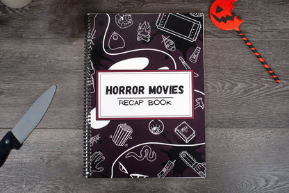 top down view of a spiral movie review journal titled Horror Movies Recap Book on wood table with pumpkin lollipop and Halloween decorations