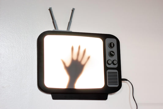 A black vintage-style television wall lamp with a light-up screen showing a shadowy hand. The lamp has a retro design with knobs and antenna details. The overall aesthetic is dark and mysterious, suitable for a horror or suspense theme. The television is black with gray details. The light from the screen is a soft white.