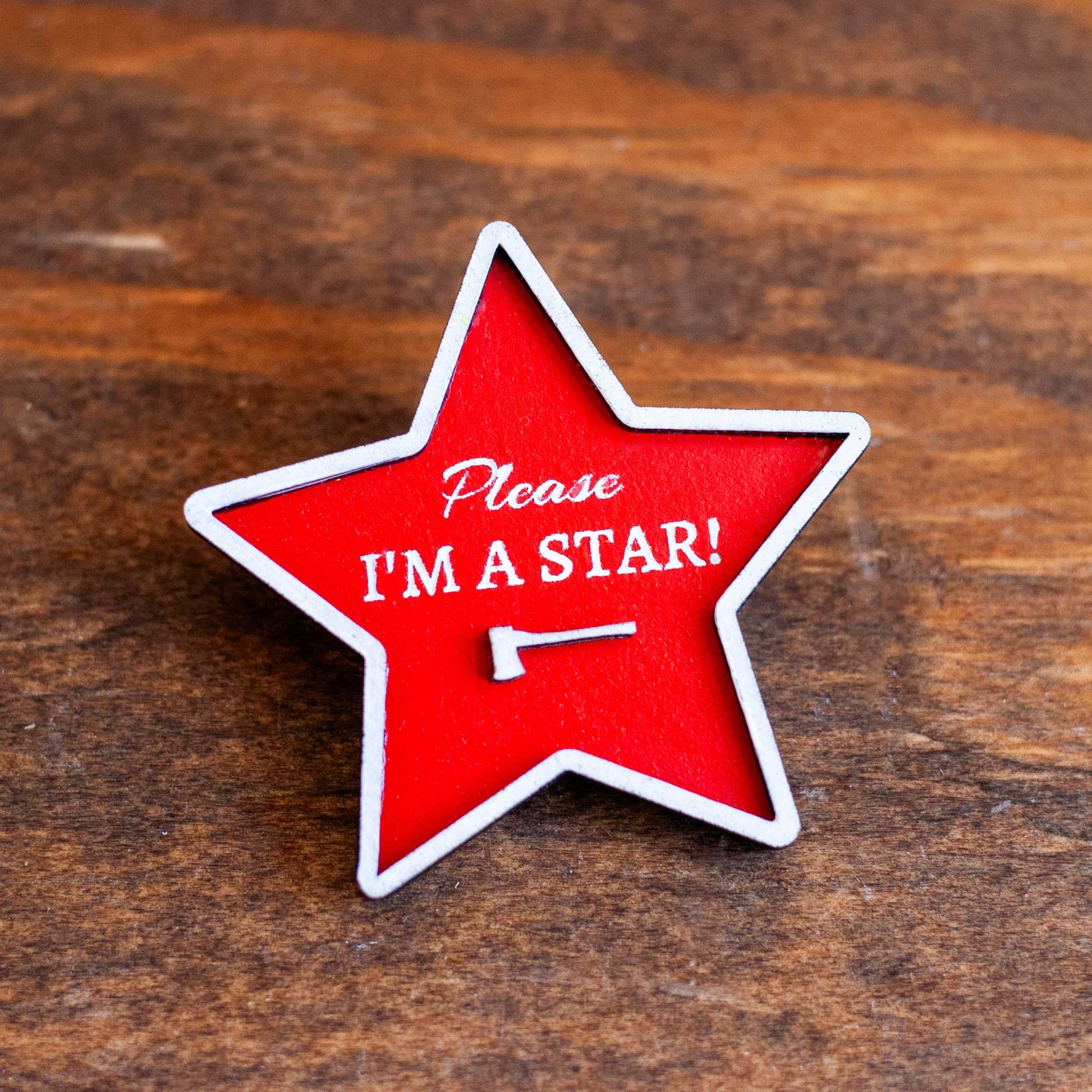 Red star-shaped brooch pin with 'Please I'm a Star!' text on a wooden surface
