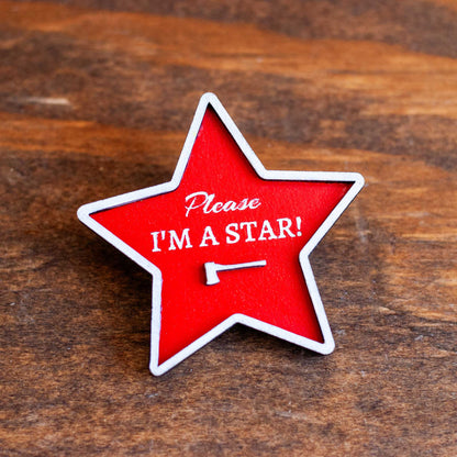 Red star-shaped brooch pin with 'Please I'm a Star!' text on a wooden surface