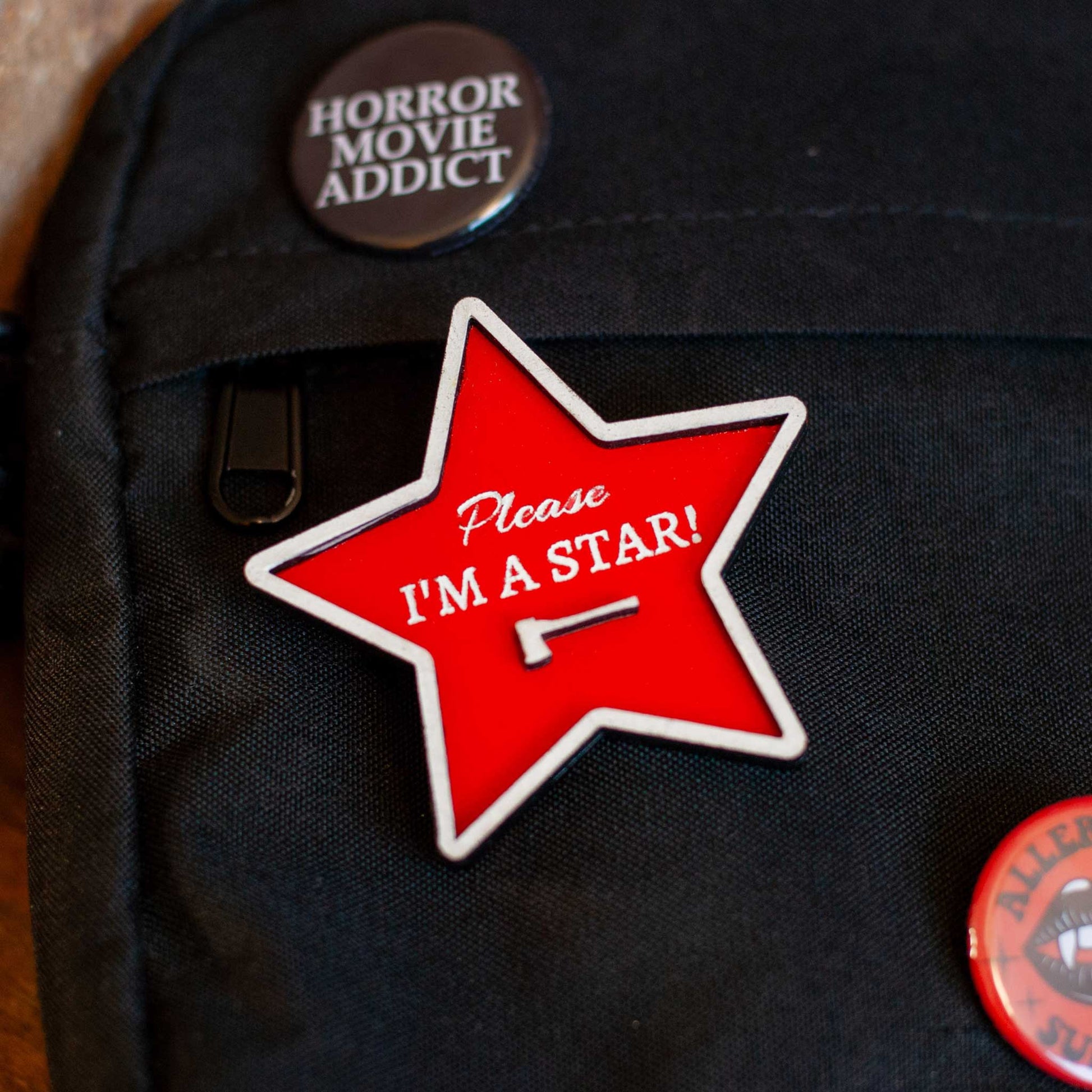 Red star-shaped pin with 'Please I'm a Star!' text on a cross-body bag with other horror pins