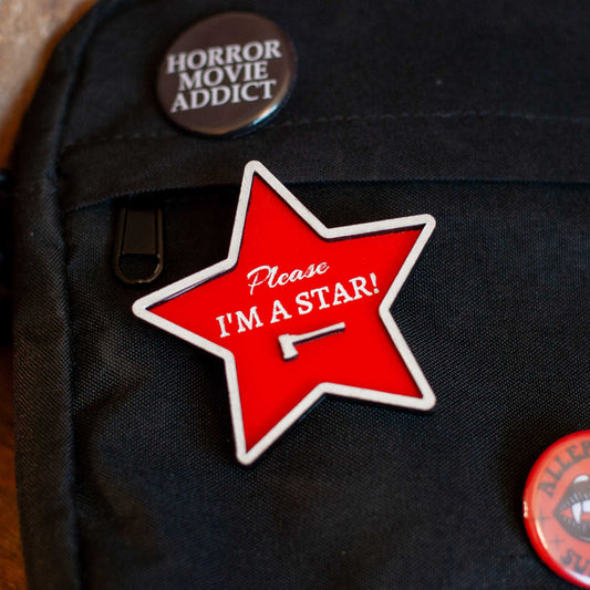 Red star-shaped pin with 'Please I'm a Star!' text on a cross-body bag with other horror pins