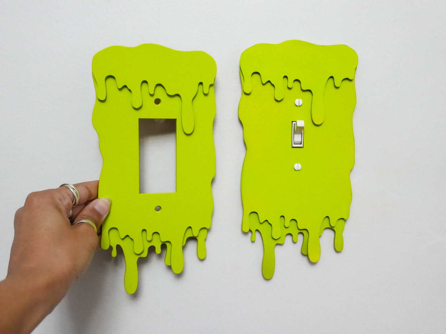 Two slime green light switch plates with a dripping design in Rocker and Toggle style held by a hand against a white background.