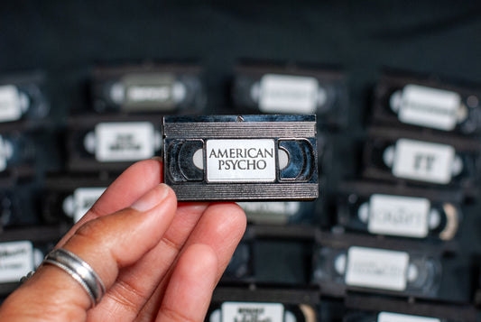Hand holding a mini VHS tape pin brooch labeled 'American Psycho' with more VHS tapes in the background.