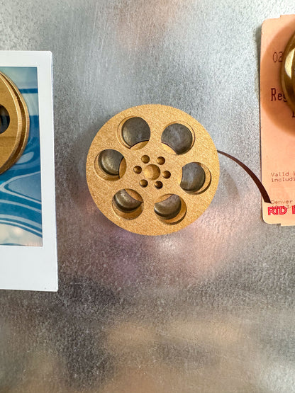 front view of a gold miniature movie reel magnet holding up a polaroid picture on fridge