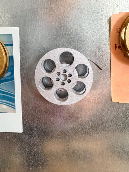 front view of a silver miniature movie reel magnet holding up a movie ticket on fridge