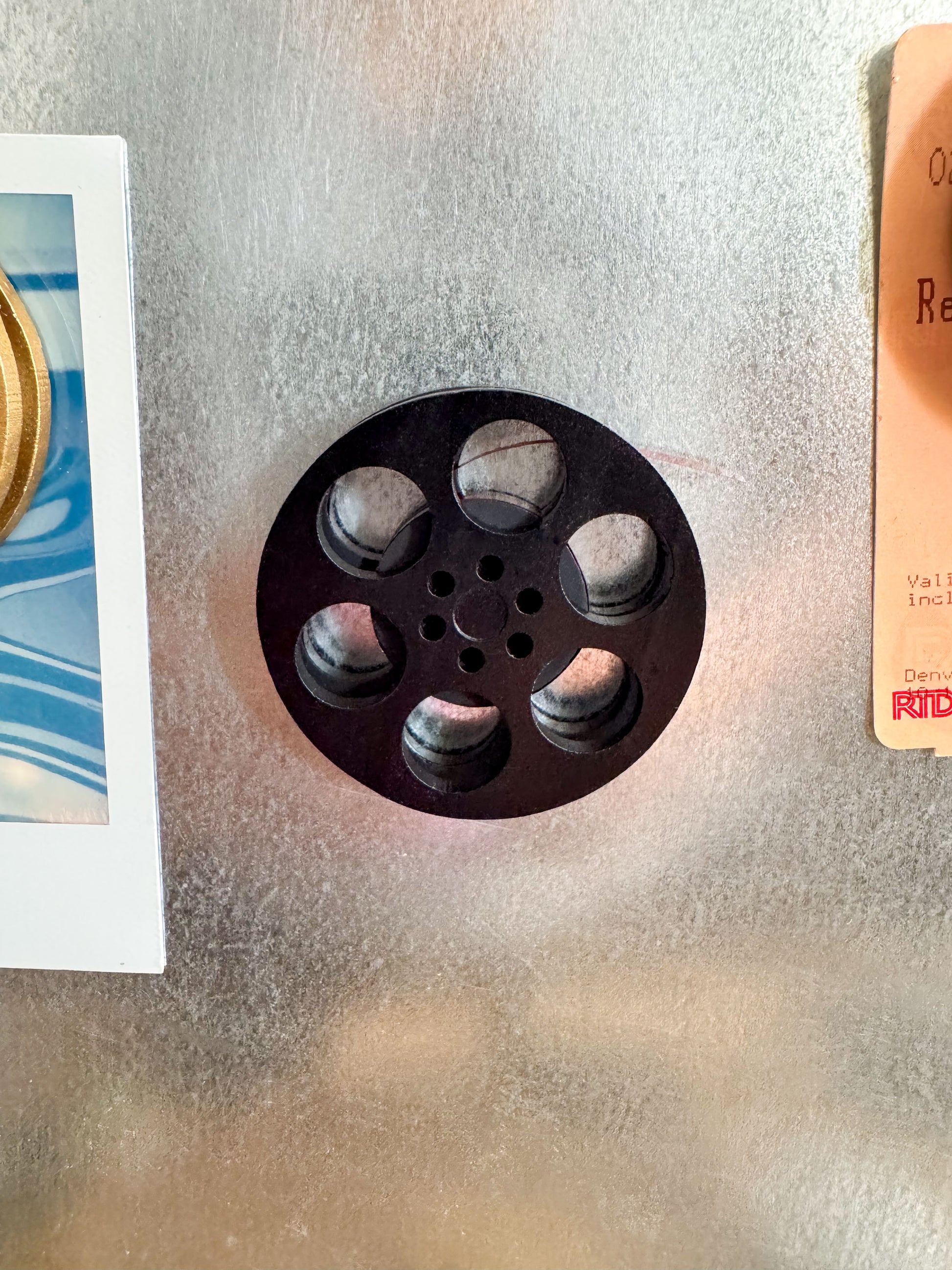 front view of a black miniature movie reel magnet holding up a polaroid picture on fridge
