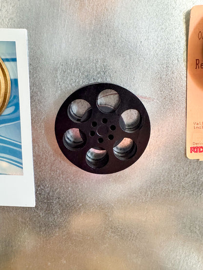 front view of a black miniature movie reel magnet holding up a polaroid picture on fridge