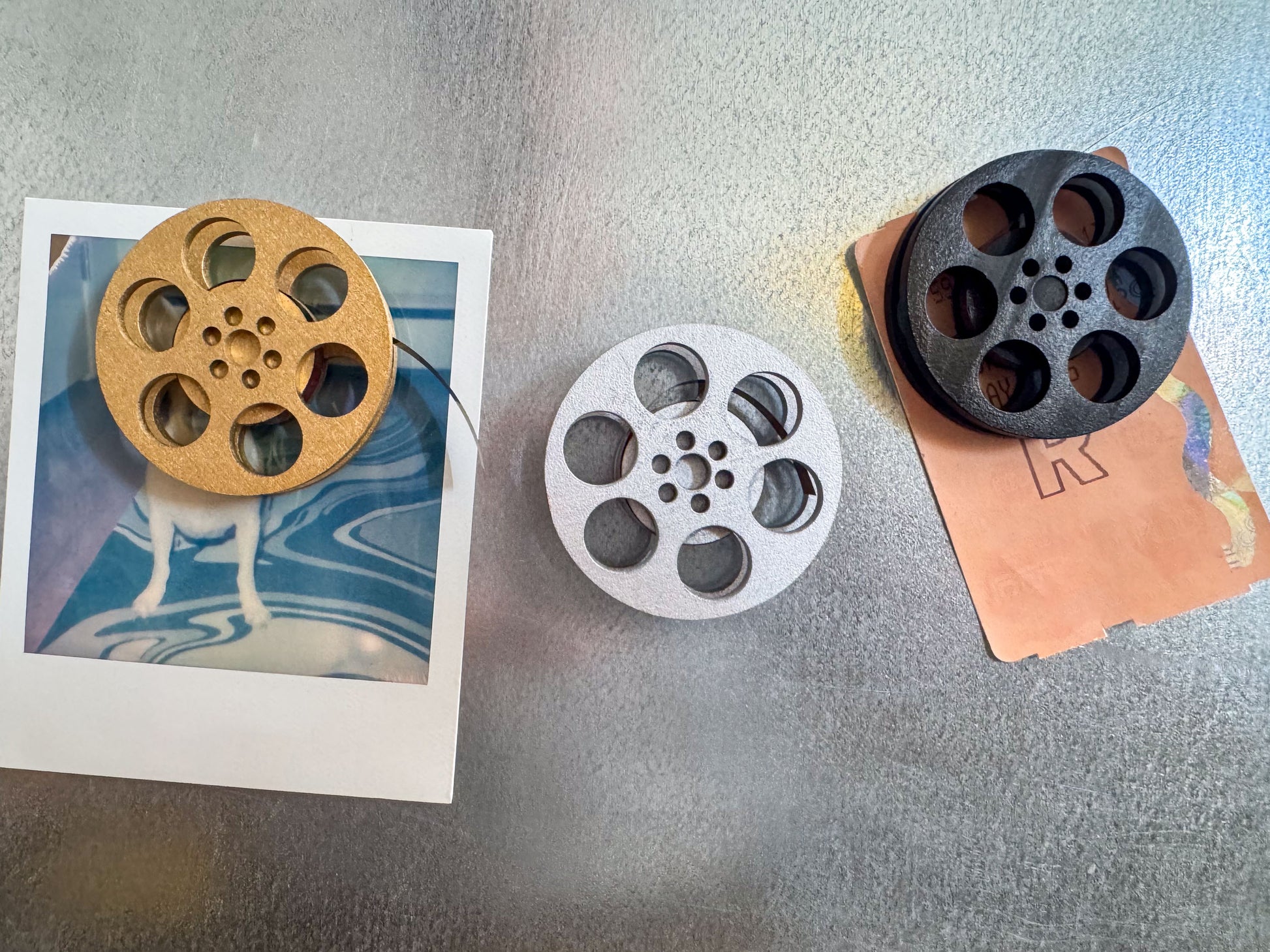 front view of three miniature movie reel magnets in gold silver and black holding up a movie ticket on fridge