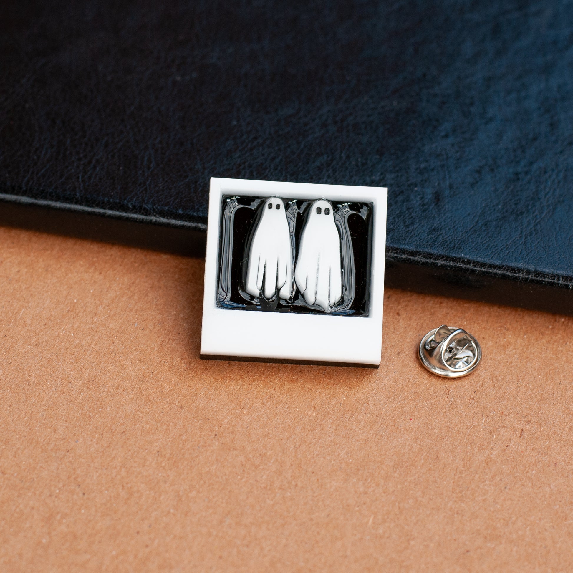 A shiny lapel pin with two white ghosts in a polaroid with no feet with silver clasp pinback next to it on wood background with black leather book in background