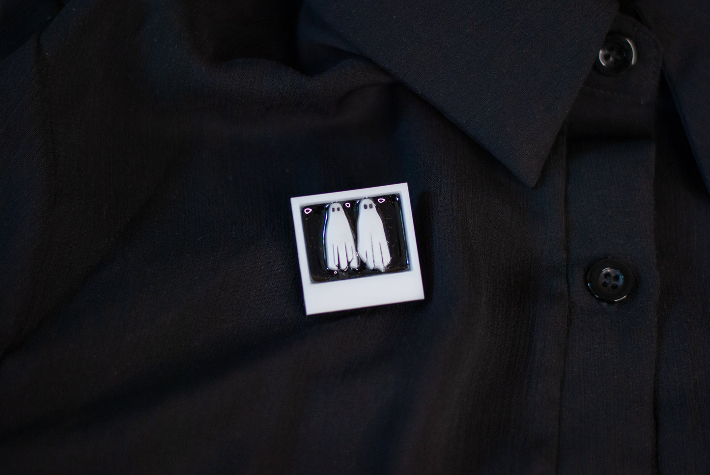 Black shirt with a white graphic pin featuring two ghosts in a white polaroid on a dark background
