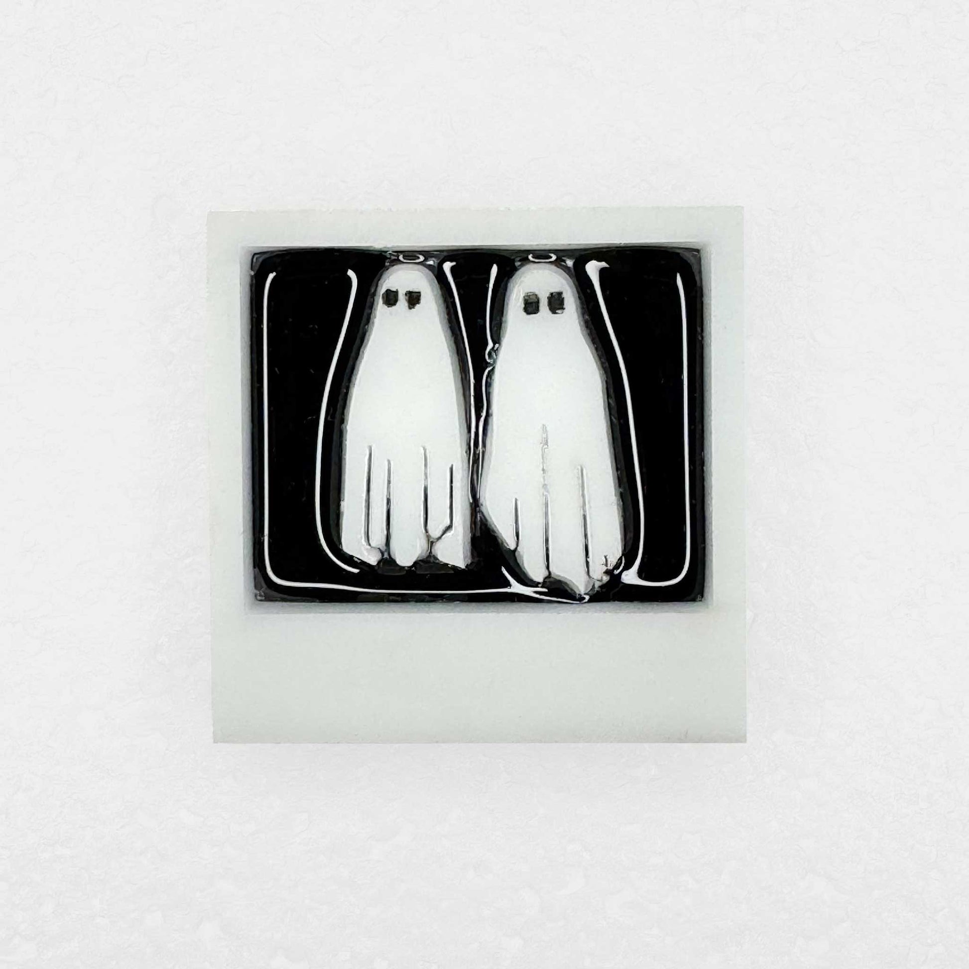 A shiny lapel pin with two white ghosts in a polaroid with no feet on white background