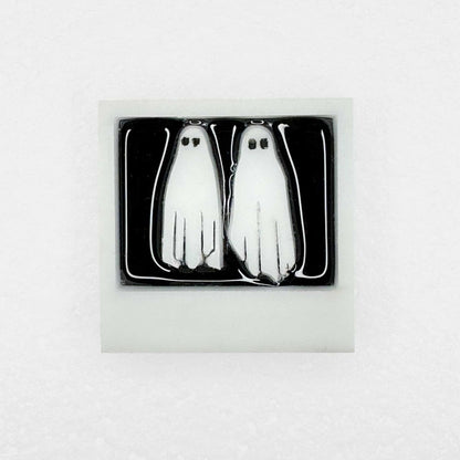 A shiny lapel pin with two white ghosts in a polaroid with no feet on white background
