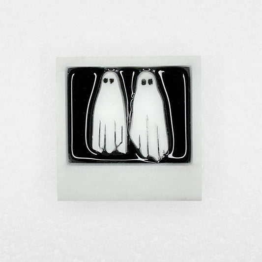 A shiny lapel pin with two white ghosts in a polaroid with no feet on white background
