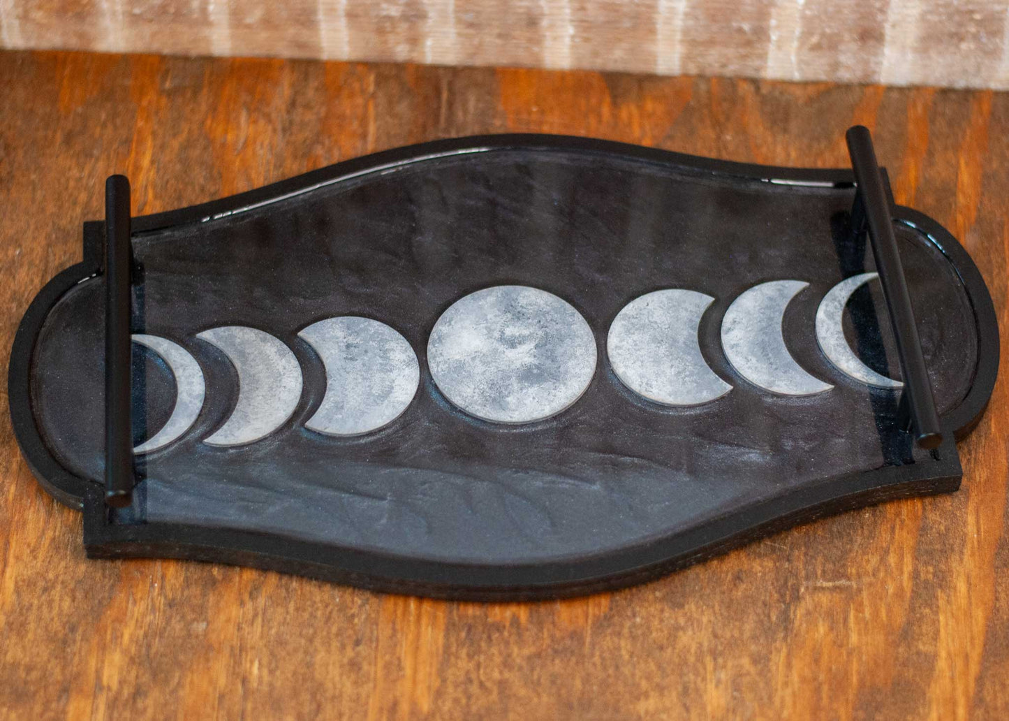 Decorative black and shimmery resin tray with moon phases with black metal handles on a wooden surface
