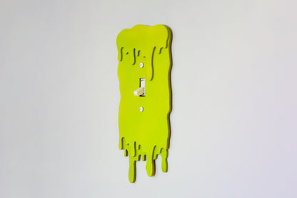 Side view of a green slime drip light switch cover plate on a white wall background