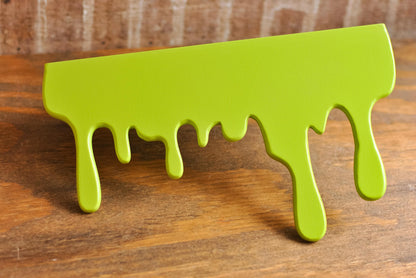 Front view of a green Slime Drip Shelf decor sitting on a wooden floating shelf, 90s Nostalgic shelf sitter, displaying Spooky Season vibes, an Accessory for Bookshelf, Display Shelf, or Mantel, Goosebumps inspired decor