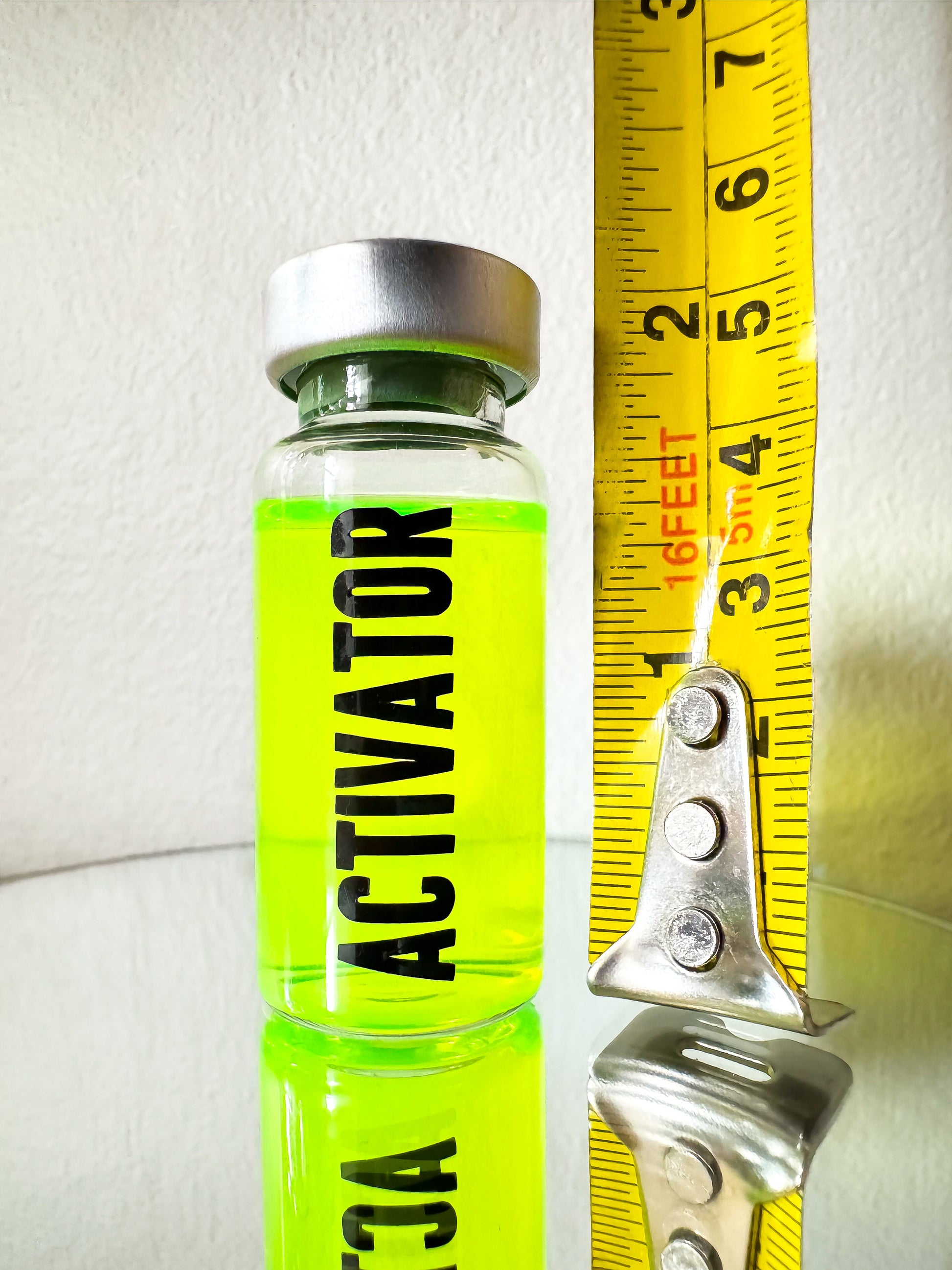 The Substance glass vial movie props labeled 'ACTIVATOR' with a green liquid next to measuring tape in front of a white background.