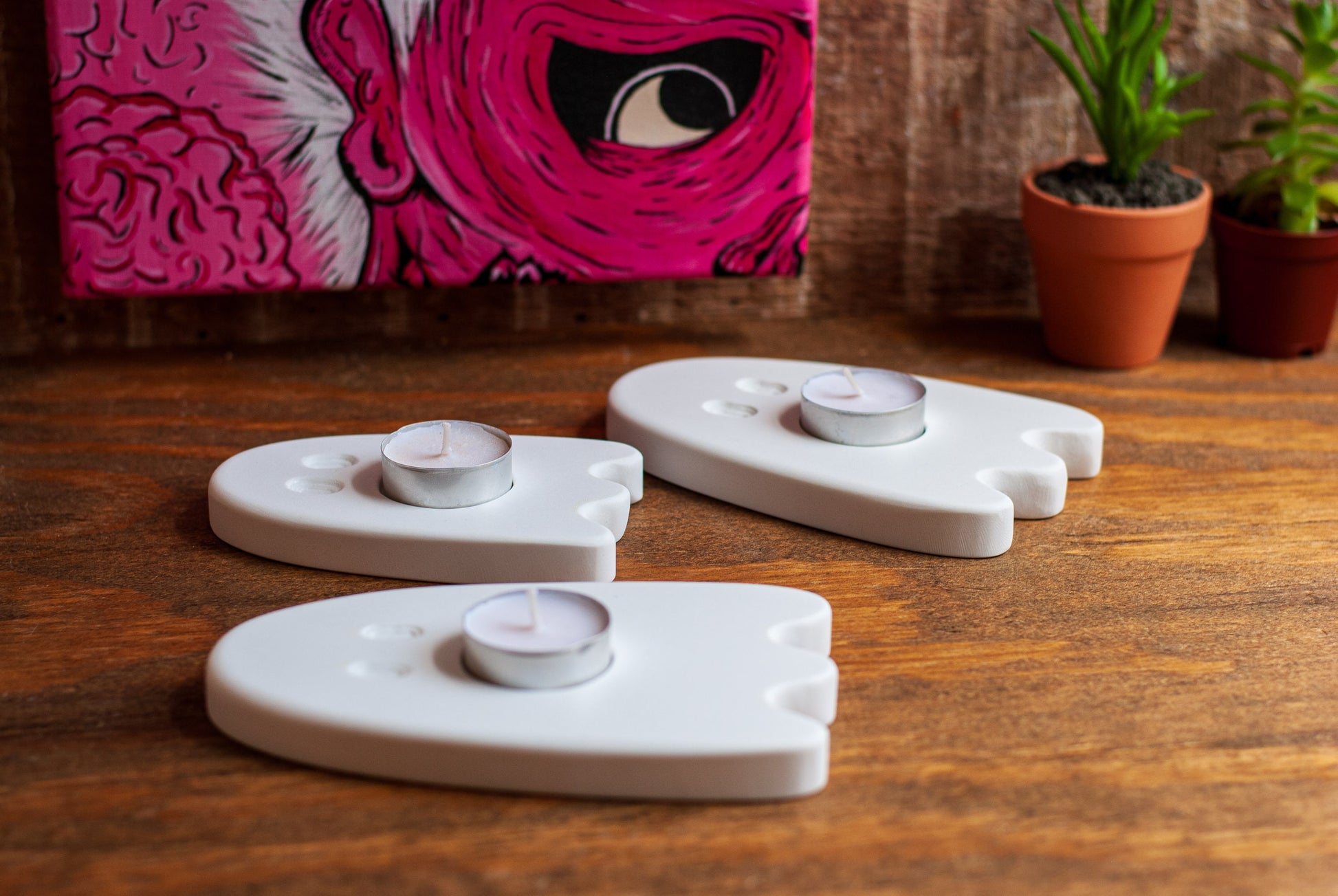 Three white ceramic ghost-shaped candle holders on a wooden surface with a pink abstract painting in the background.