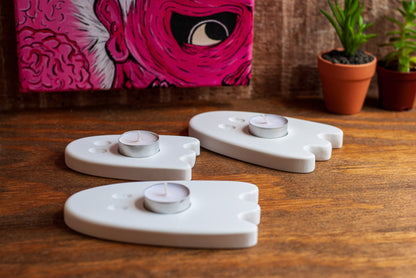 Three white ceramic ghost-shaped candle holders on a wooden surface with a pink abstract painting in the background.