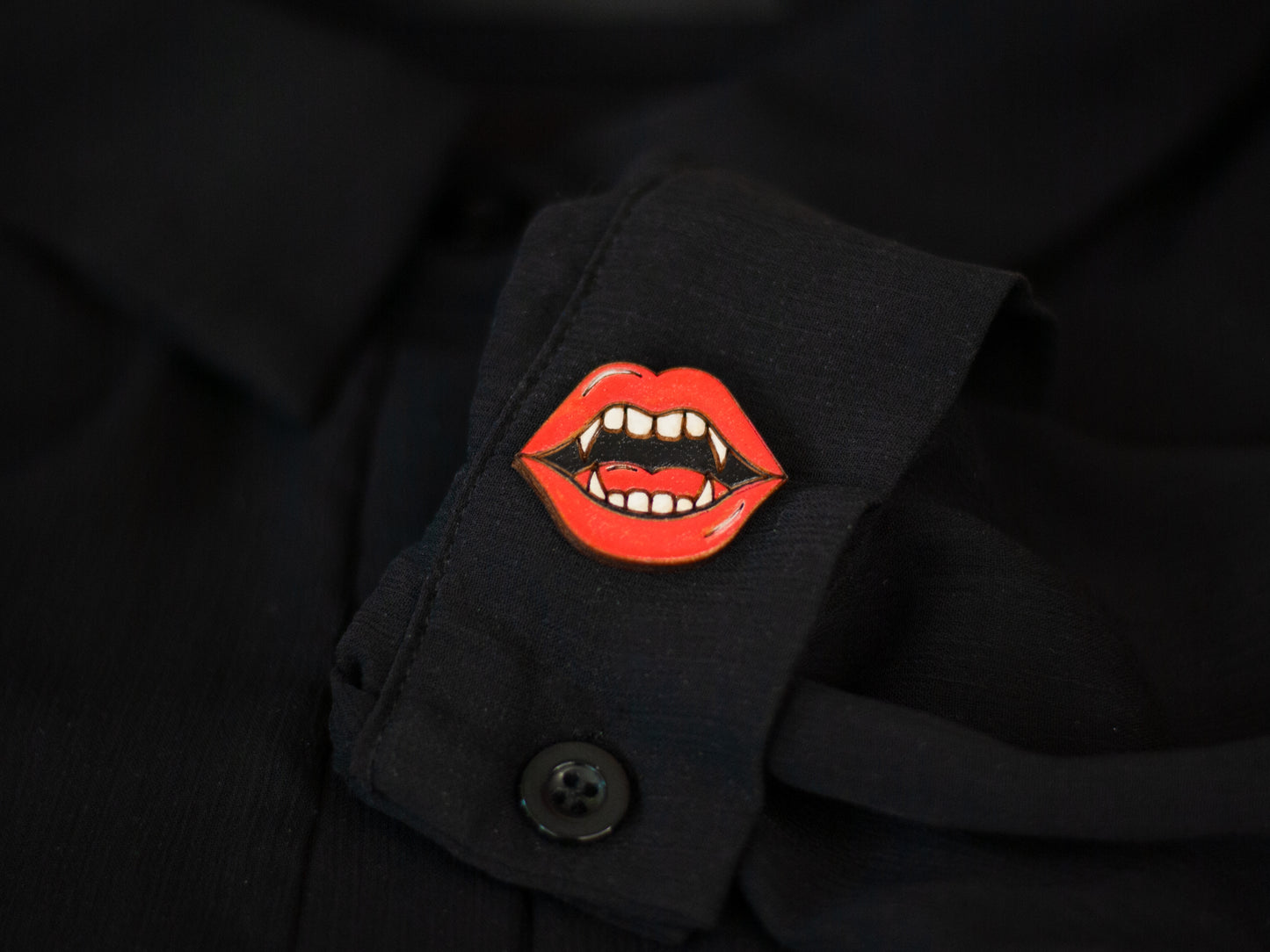 a lapel pin shaped like a vampire mouth with bright red lips and fangs to be used for book bag, shirt, or purse on a black sheer shirt cuff sleeve