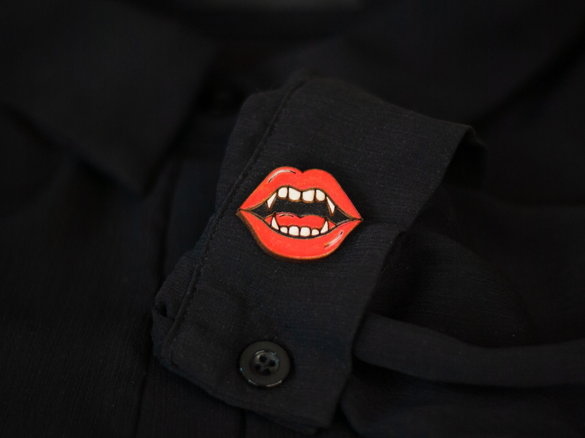 a lapel pin shaped like a vampire mouth with bright red lips and fangs to be used for book bag, shirt, or purse on a black sheer shirt cuff sleeve