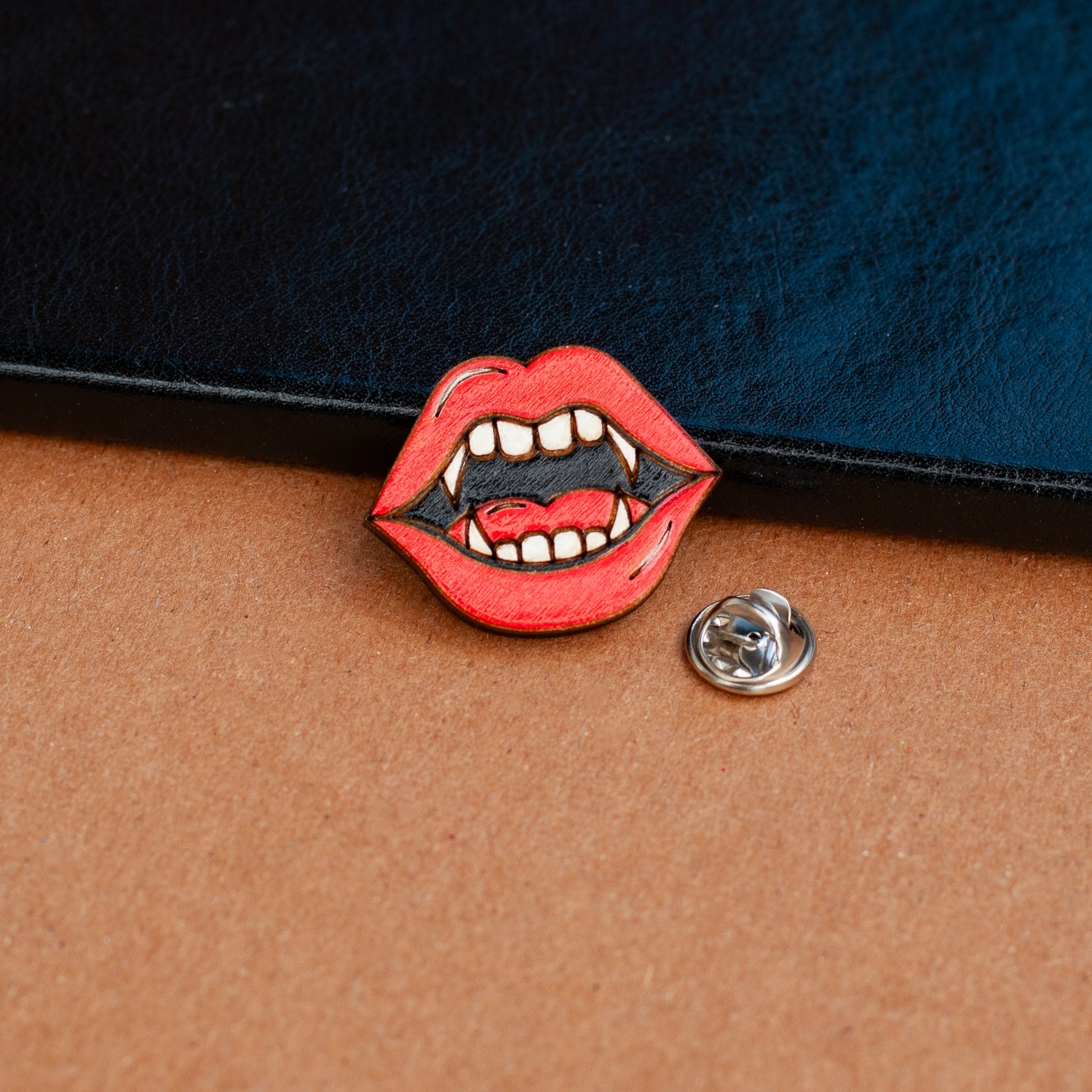 a lapel pin shaped like a vampire mouth with bright red lips and fangs and silver clasp pinback next to it. To be used for book bag, shirt, or purse on a wood background with leather book in background