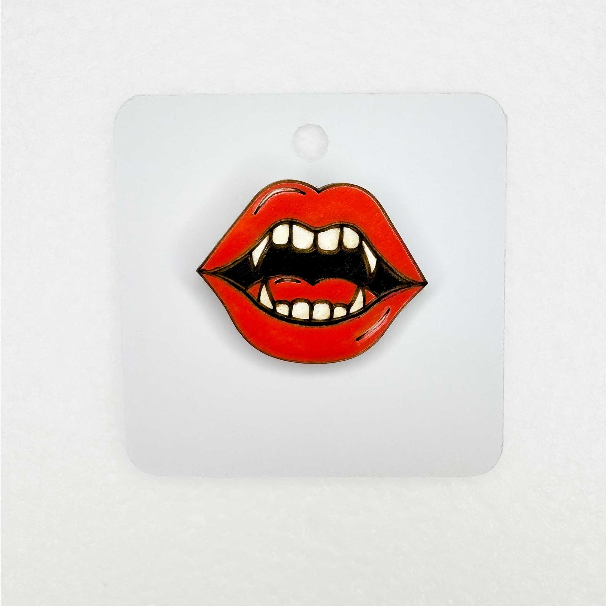 Lip-shaped lapel pin accessory with vampire teeth design on a white backing card with white background 